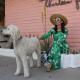 A woman wearing a green dress sits outside of a pink building in Palm Springs with a white dog