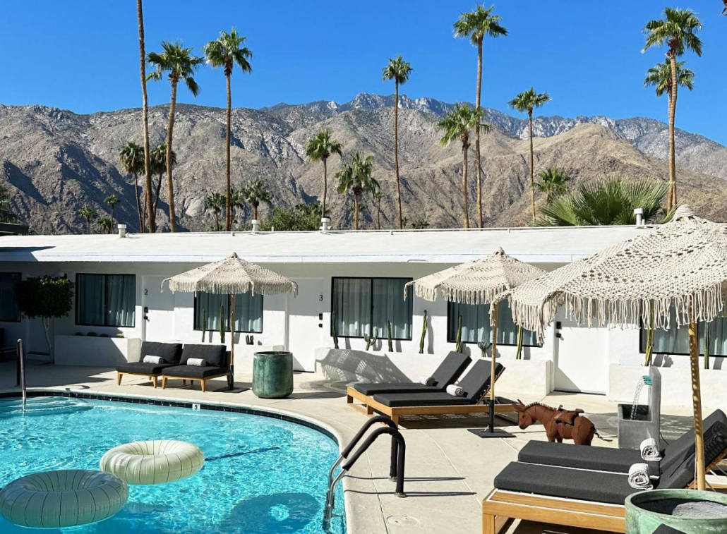 The pool at Jazz Hotel surrounded by fringed umbrellas and lounge chairs with a white ring float in the water and palms and San Jacinto Mountains in the background