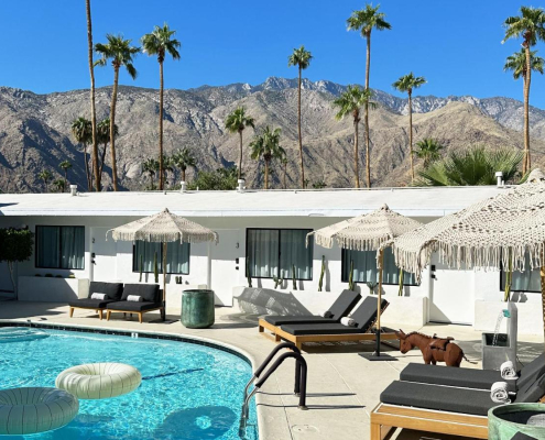 The pool at Jazz Hotel surrounded by fringed umbrellas and lounge chairs with a white ring float in the water and palms and San Jacinto Mountains in the background