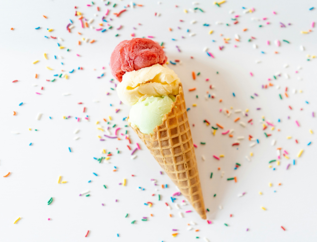 A three-scoop ice cream cone on a white background surrounded by colorful rainbow sprinkles