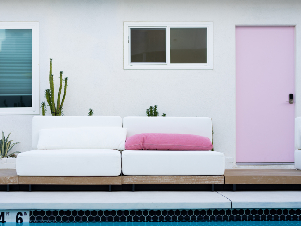 A white outdoor couch with pink pillows in front of a pink door and white walls in the courtyard of The Cactai