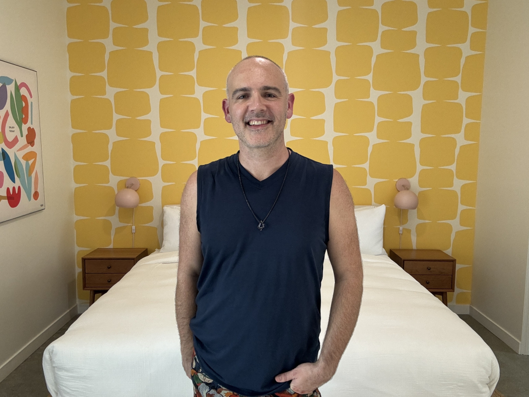 A white man stands in front of a yellow wallpapered bedroom at The Cactai Palm Springs