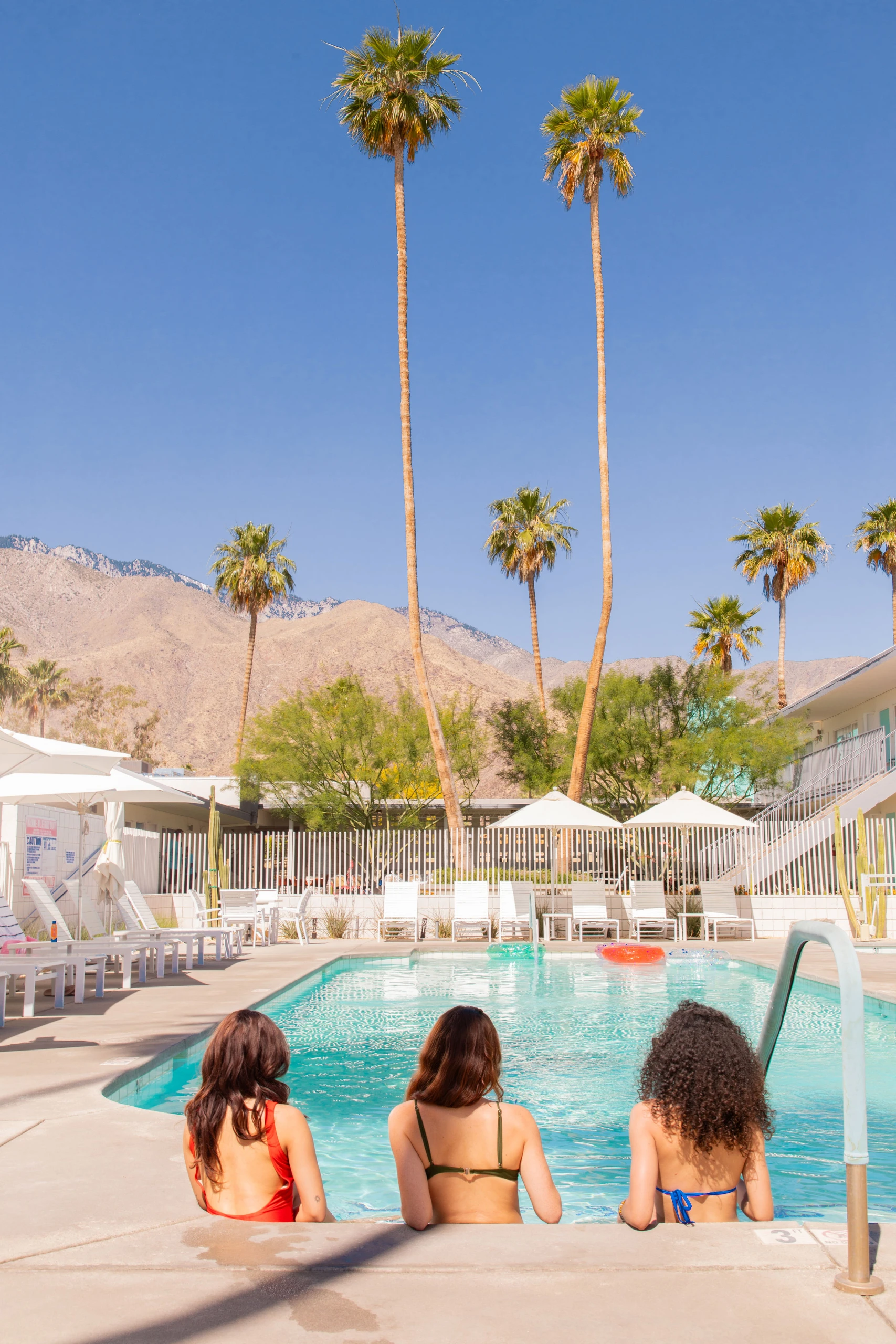Three women wearing bikinis sit in the Skylark Hotel pool with their back turned to the camera facing palm trees
