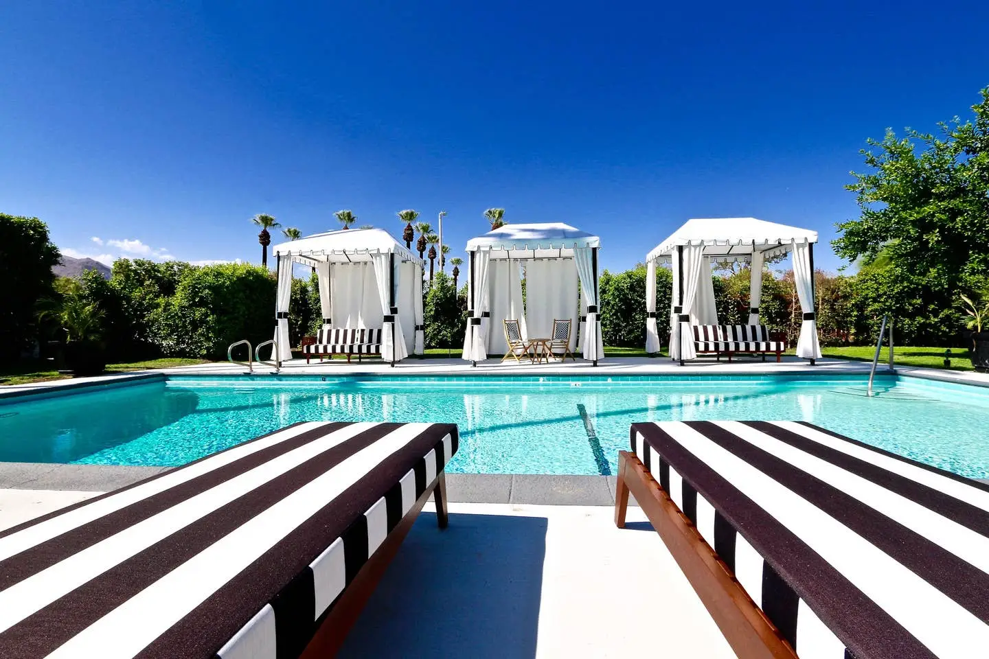 Black and white striped lounge chairs in front of the pool and next to three outdoor cabanas at Hotel El Cid in Palm Springs, California