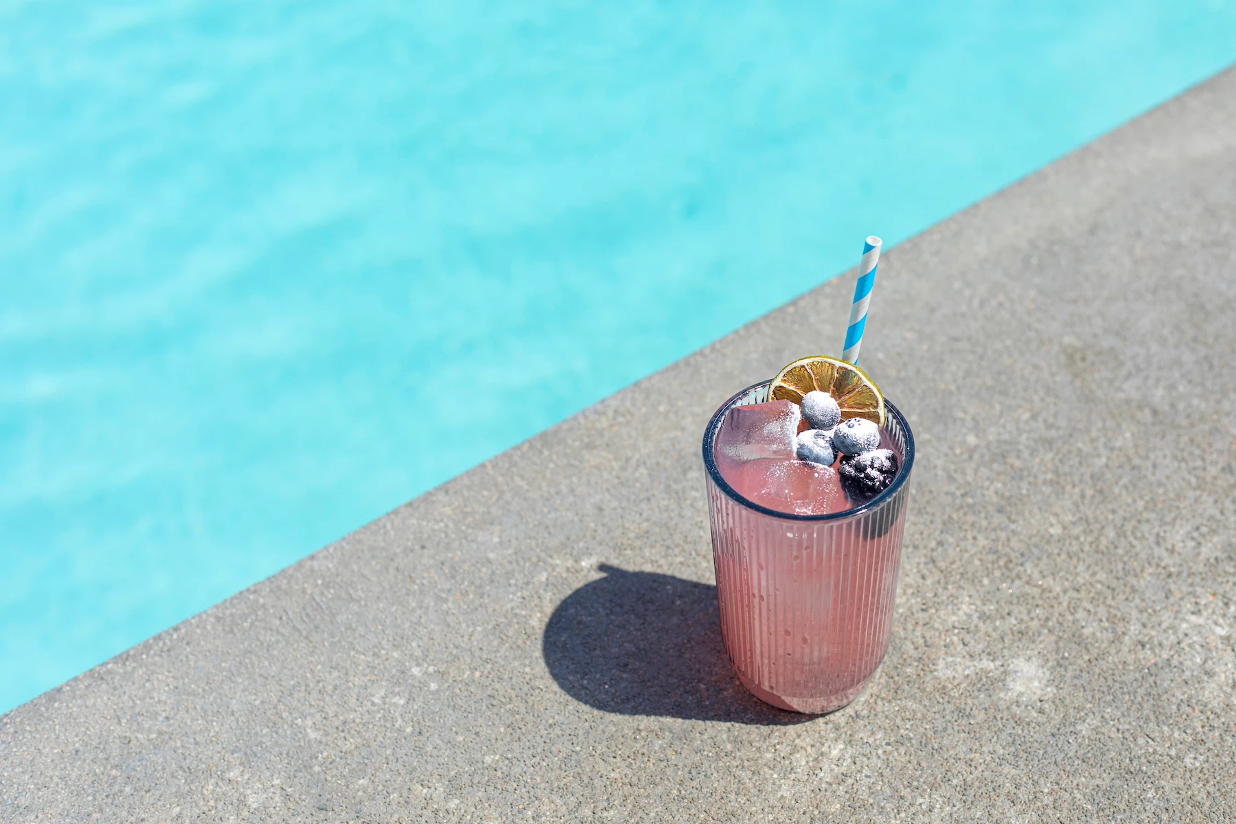 A glass holding a pink drink topped with blueberries next to a pool