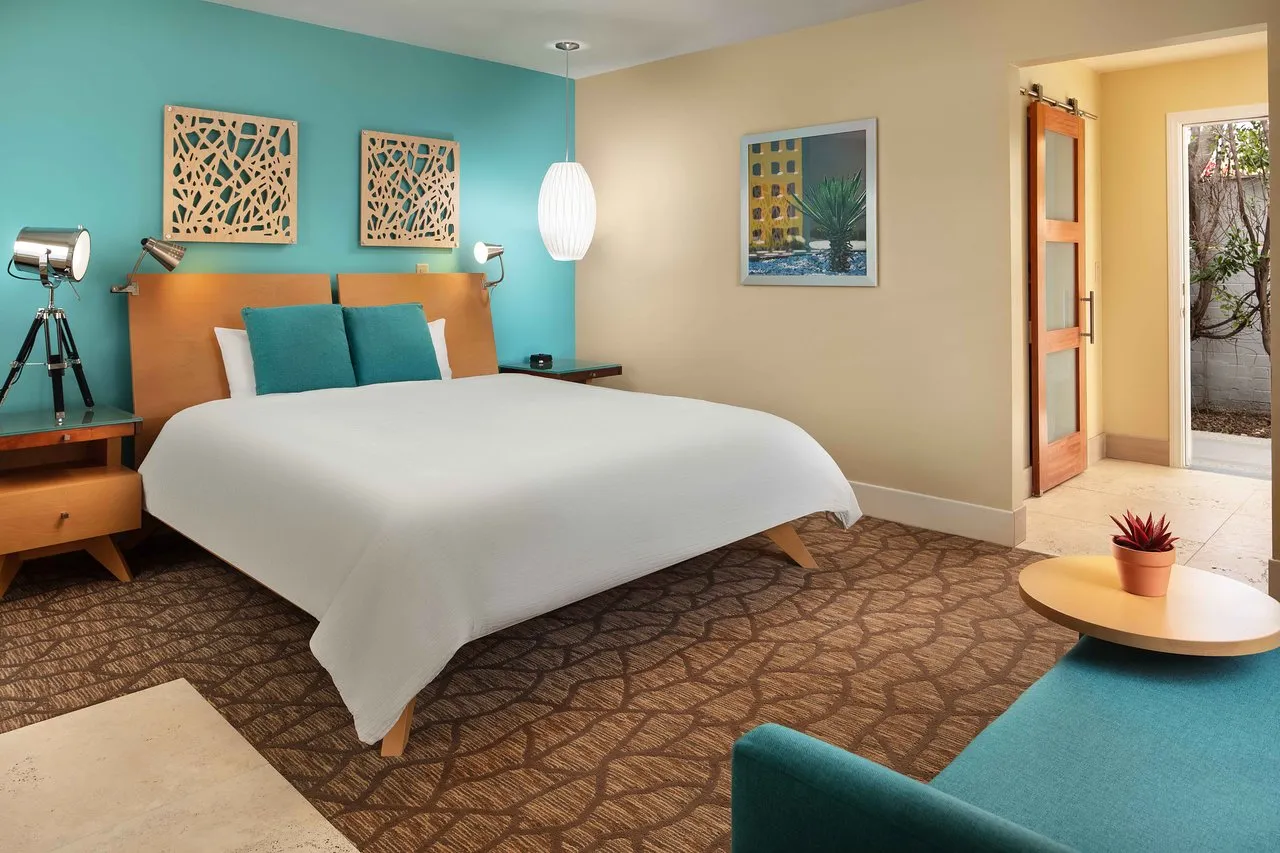 A bedroom at Movie Colony Hotel in Palm Springs, California, with a bed topped with a white comforter and blue pillows and a blue couch with modernist art on the walls