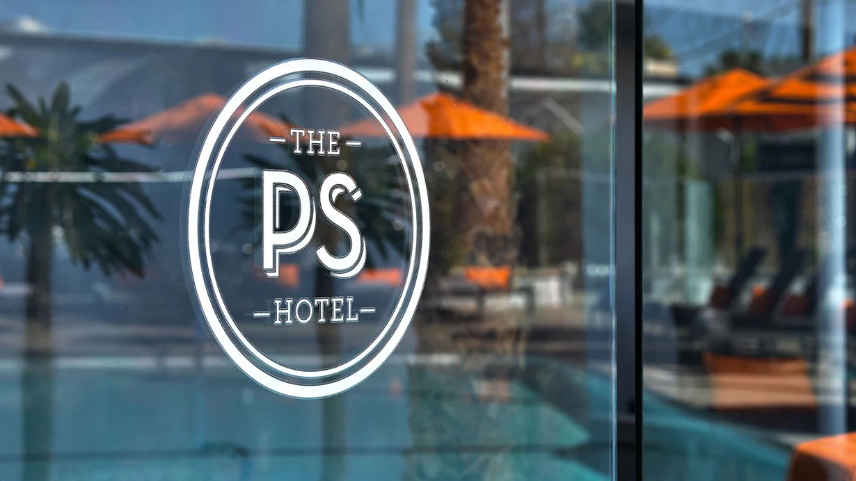 A window at The Palm Springs Hotel with "The PS Hotel" on a decal with orange umbrellas reflected off of it