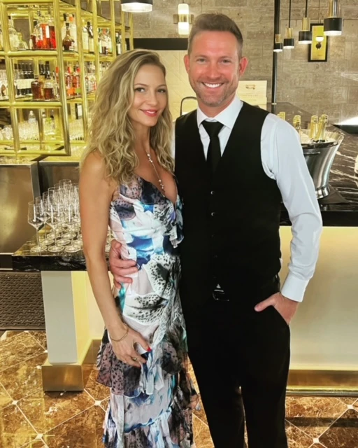 A blonde woman in a white dress and white man in a dress vest and pants stand in a restaurant with the bar behind them