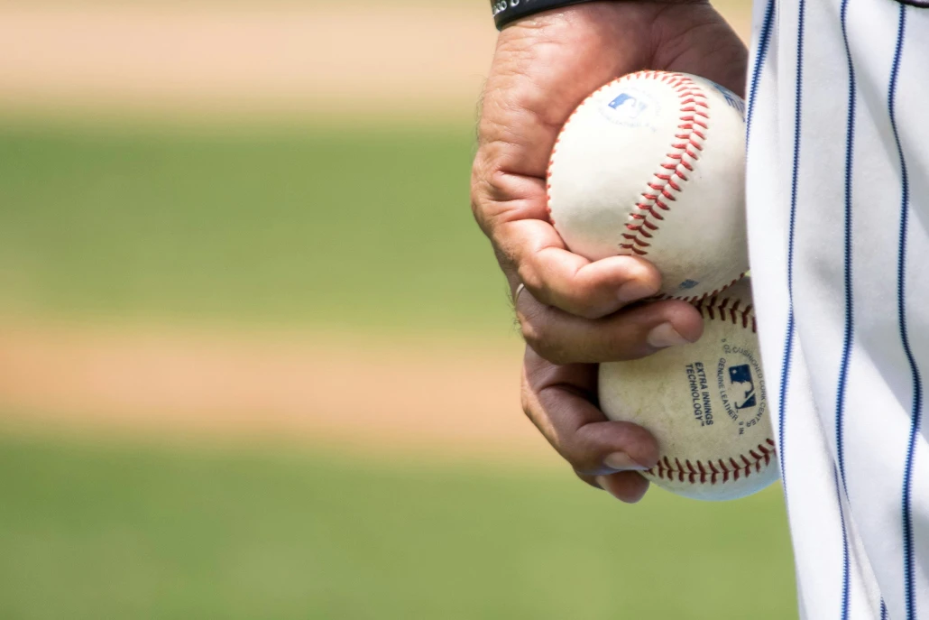 A man in a baseball uniform on the field holds two baseballs in his left hand