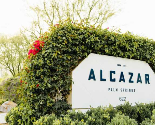 A white sign that says Alcazar Palm Springs in blue letters surrounded by green shrubbery